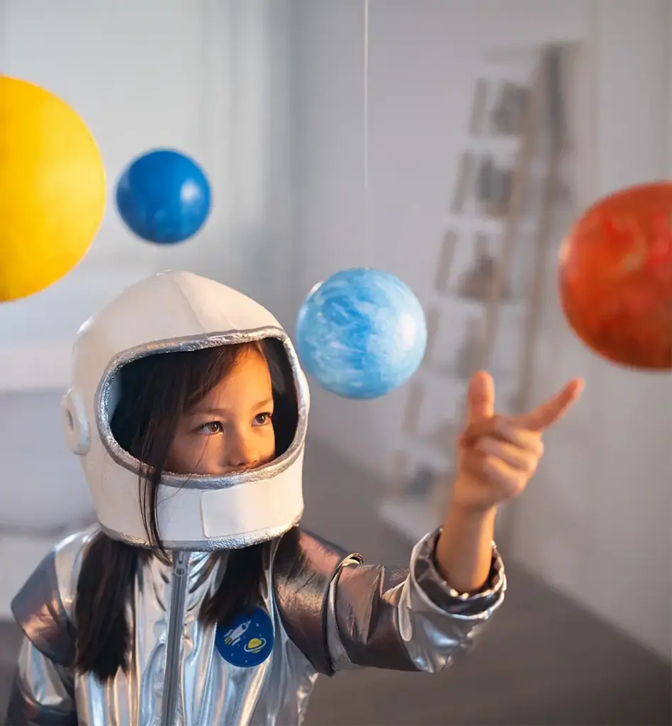 Kleines Mädchen im silbernen Astronauten-Kostüm zeigt begeistert auf bunte, von der Decke hängende Planeten-Modelle.