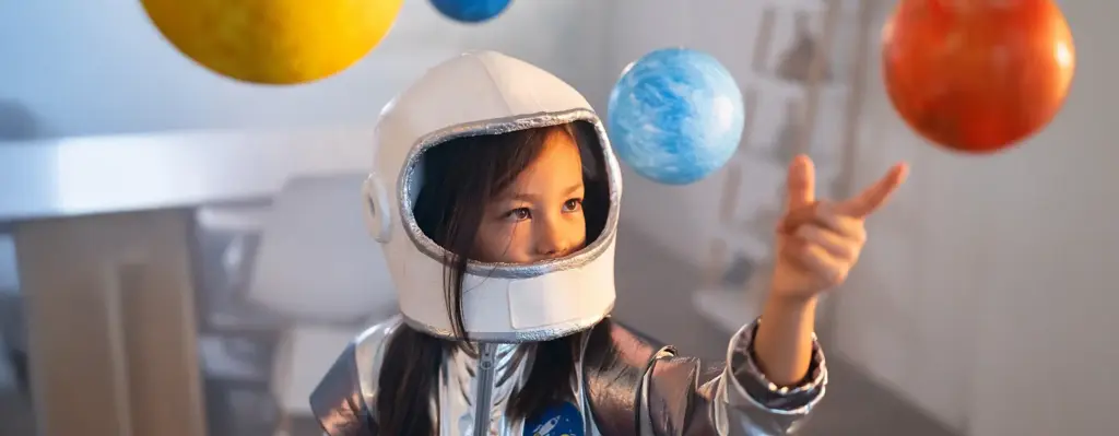 Kleines Mädchen im silbernen Astronauten-Kostüm zeigt begeistert auf bunte, von der Decke hängende Planeten-Modelle.
