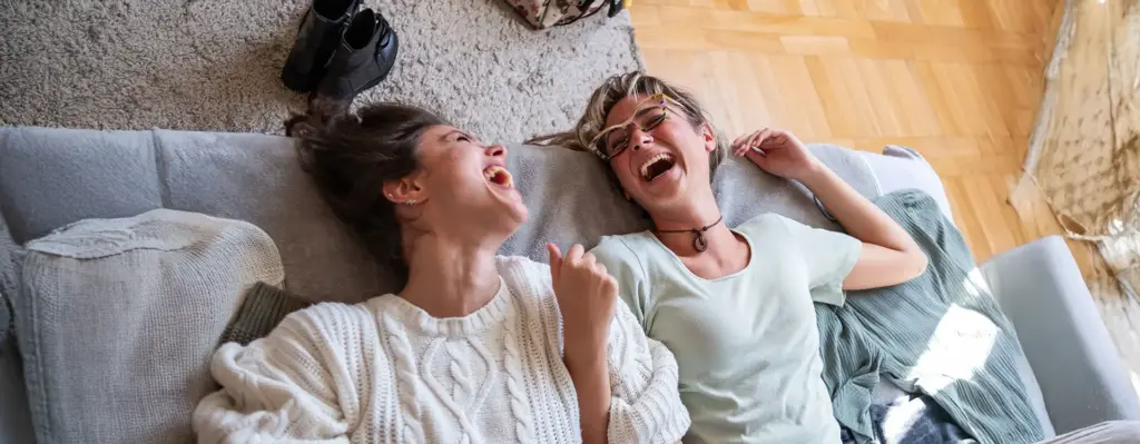 Zwei Freundinnen liegen auf einem Sofa und lachen sich ausgelassen schlapp – herzliche, unbeschwerte Stimmung zuhause.