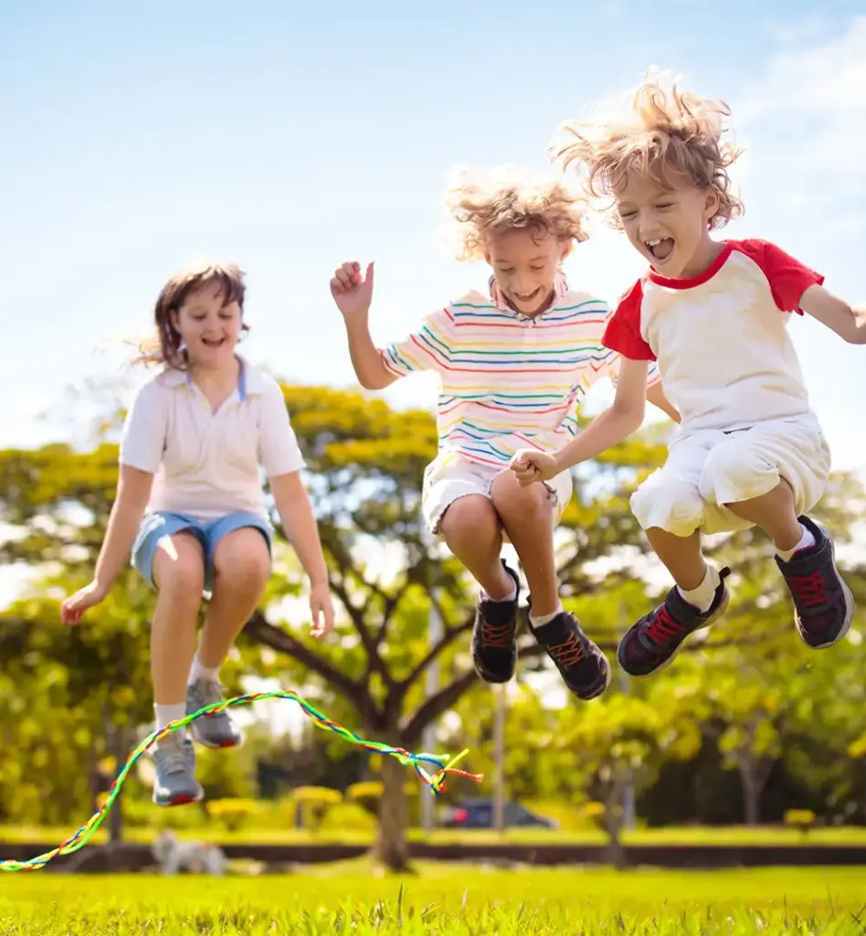 Drei Kinder springen auf einer sonnigen Wiese gemeinsam Seil und lachen – ausgelassenes Spielen im Freien an einem Sommertag.