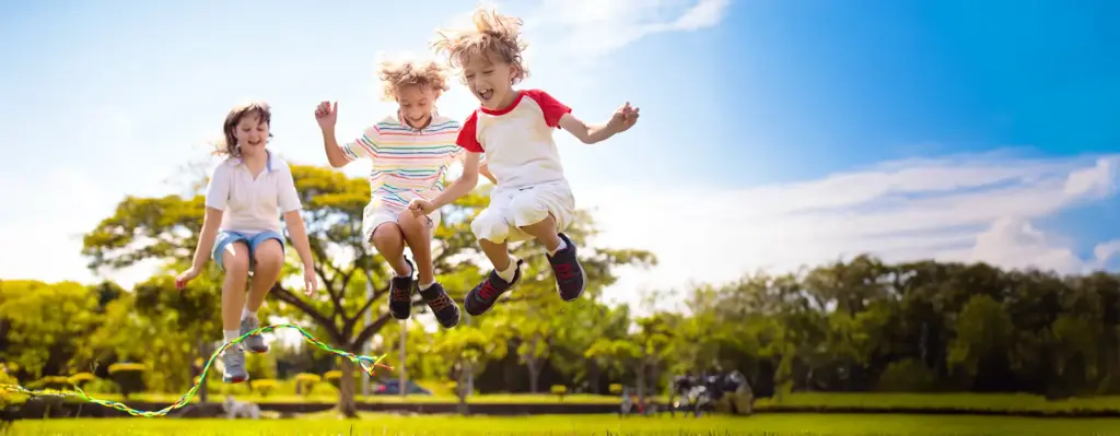 Drei Kinder springen auf einer sonnigen Wiese gemeinsam Seil und lachen – ausgelassenes Spielen im Freien an einem Sommertag.