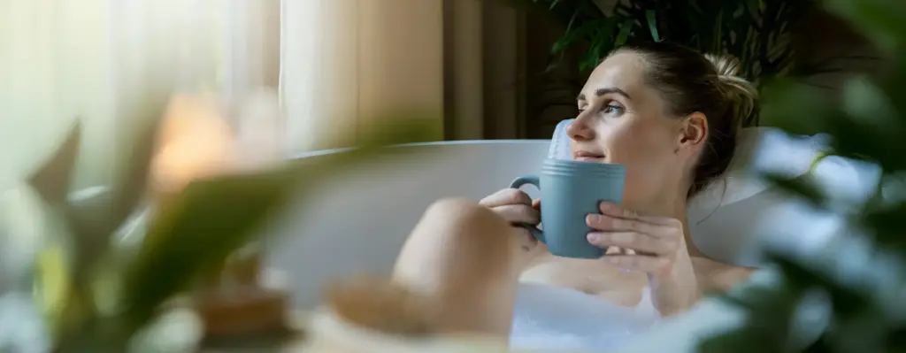 Frau entspannt in einer Schaumbadewanne, hält eine Tasse Tee und blickt zufrieden nach oben – Entspannung und Selbstfürsorge.