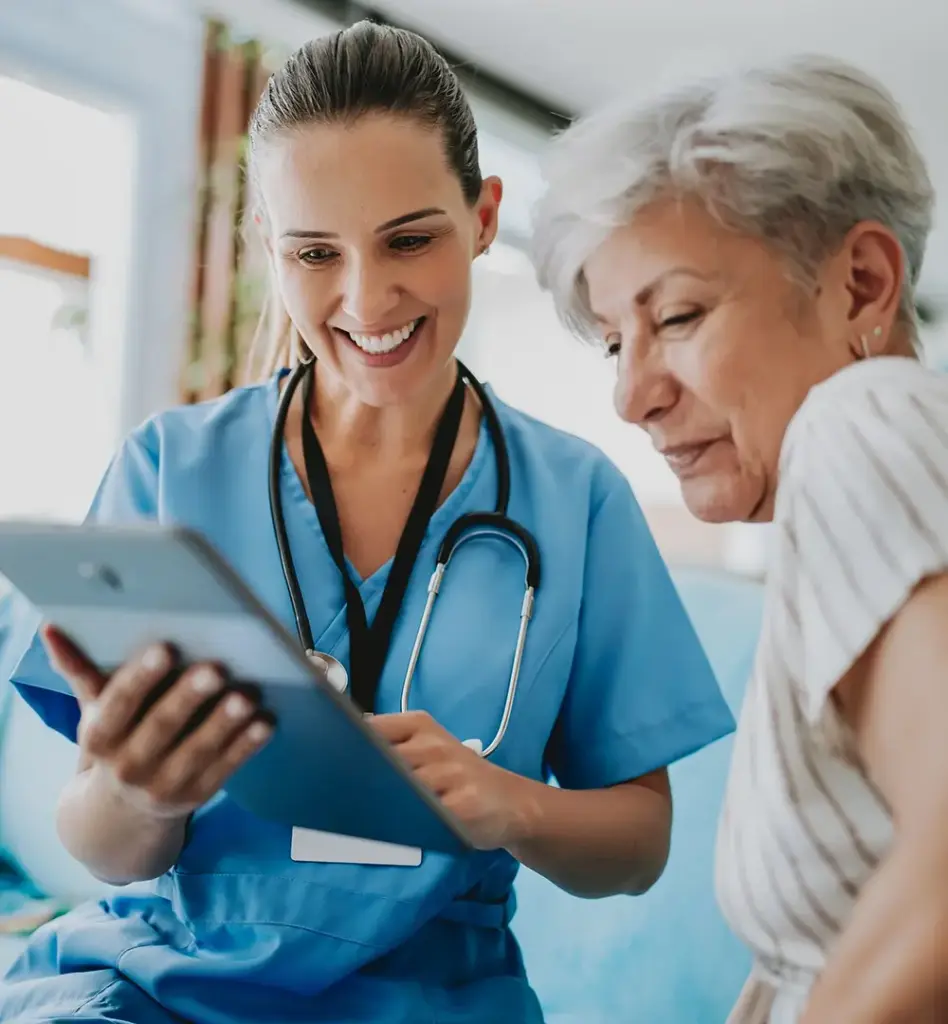 Ärztin in blauem Kasack zeigt einer älteren Patientin Gesundheitsdaten auf einem Tablet – beide lächeln, vertrauensvolle Arzt-Patientin-Situation.