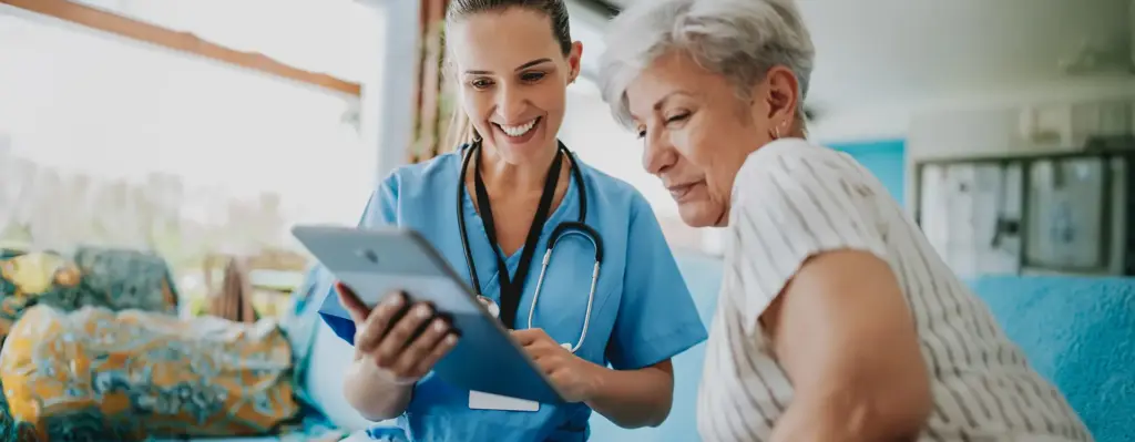 Ärztin in blauem Kasack zeigt einer älteren Patientin Gesundheitsdaten auf einem Tablet – beide lächeln, vertrauensvolle Arzt-Patientin-Situation.