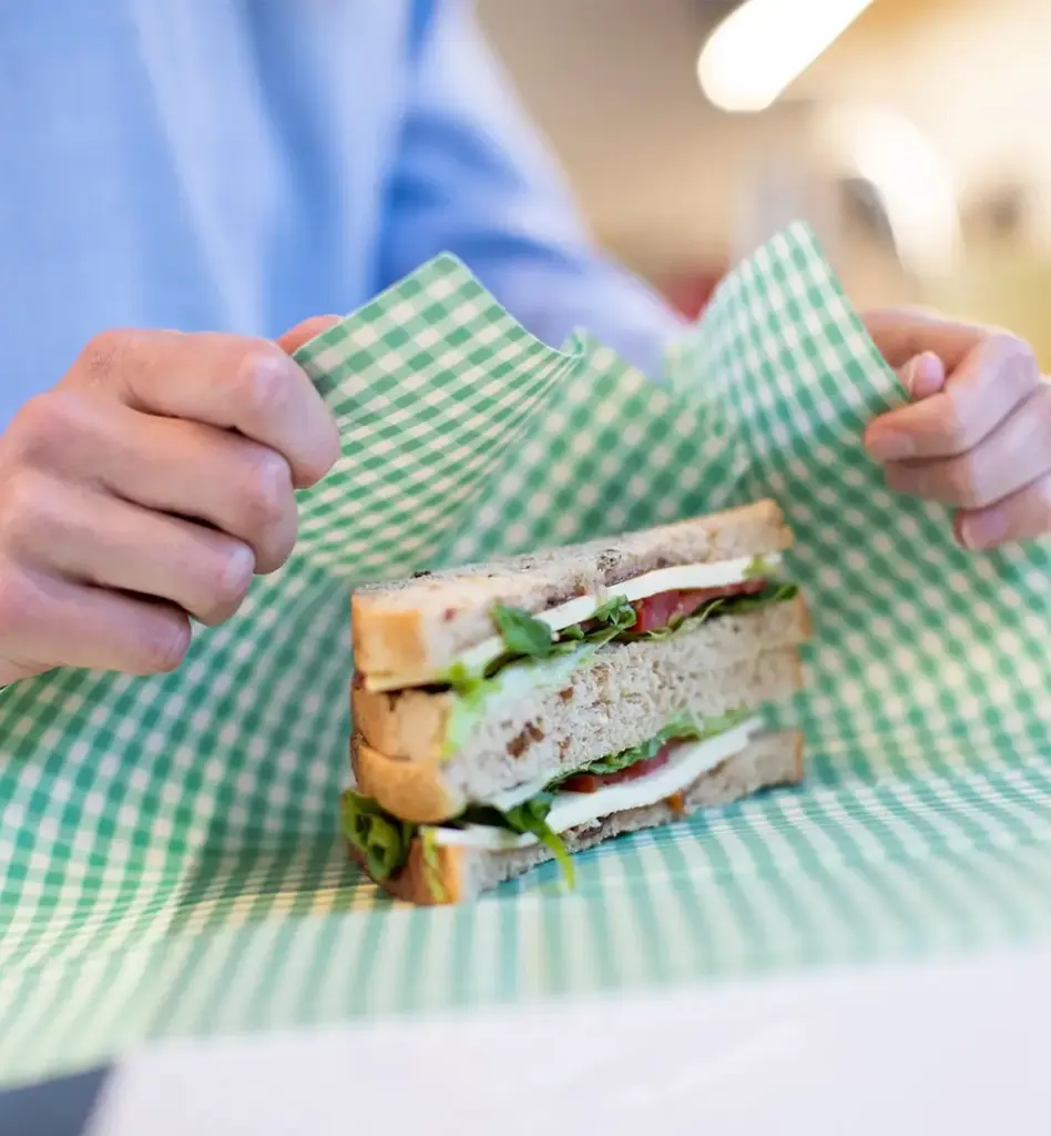 Hände wickeln ein frisches Sandwich mit Salat in ein grün-weiß kariertes Bienenwachs-Tuch – nachhaltiges und plastikfreies Einpacken.