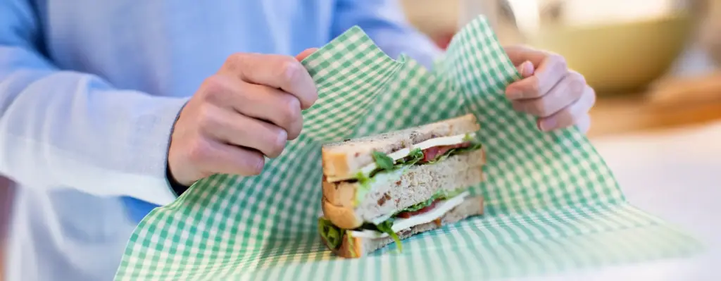Hände wickeln ein frisches Sandwich mit Salat in ein grün-weiß kariertes Bienenwachs-Tuch – nachhaltiges und plastikfreies Einpacken.