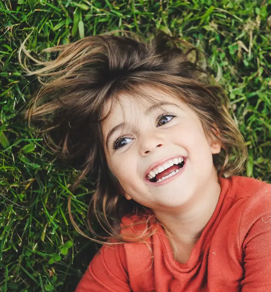 Kind liegt lachend auf einer grünen Wiese und schaut strahlend in die Kamera – ausgelassene Freude im Freien.