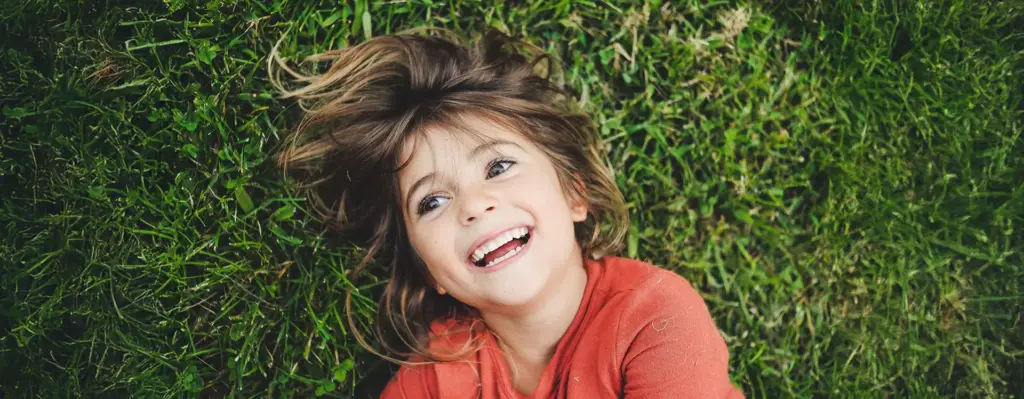 Kind liegt lachend auf einer grünen Wiese und schaut strahlend in die Kamera – ausgelassene Freude im Freien.