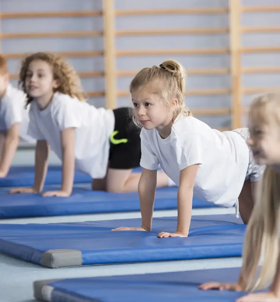 Kinder machen Liegestützen auf blauen Turnmatten in einer Sporthalle – Schulsport mit konzentriertem Gesichtsausdruck.