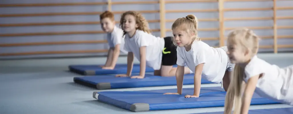 Kinder machen Liegestützen auf blauen Turnmatten in einer Sporthalle – Schulsport mit konzentriertem Gesichtsausdruck.