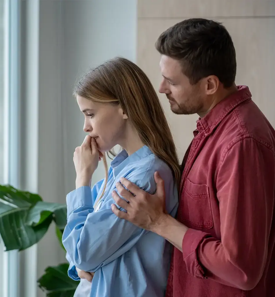 Mann legt fürsorglich die Hände auf die Schultern seiner Freundin, die besorgt aus dem Fenster schaut – emotionale Unterstützung.