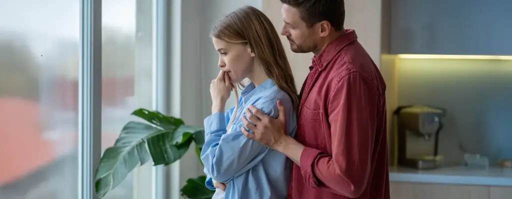 Mann legt fürsorglich die Hände auf die Schultern seiner Freundin, die besorgt aus dem Fenster schaut – emotionale Unterstützung.