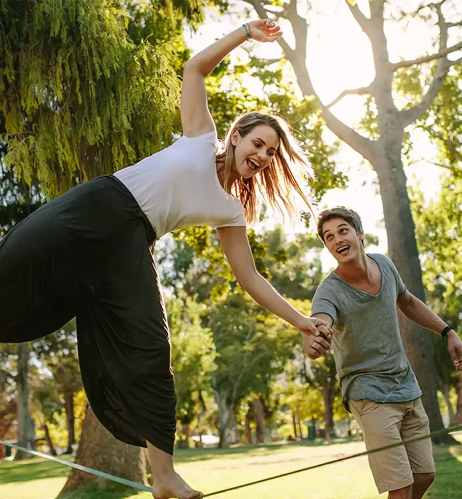 Junges Paar beim Slacklinen im Park – Frau balanciert auf dem Seil mit erhobenen Armen, Mann hält lachend ihre Hand.