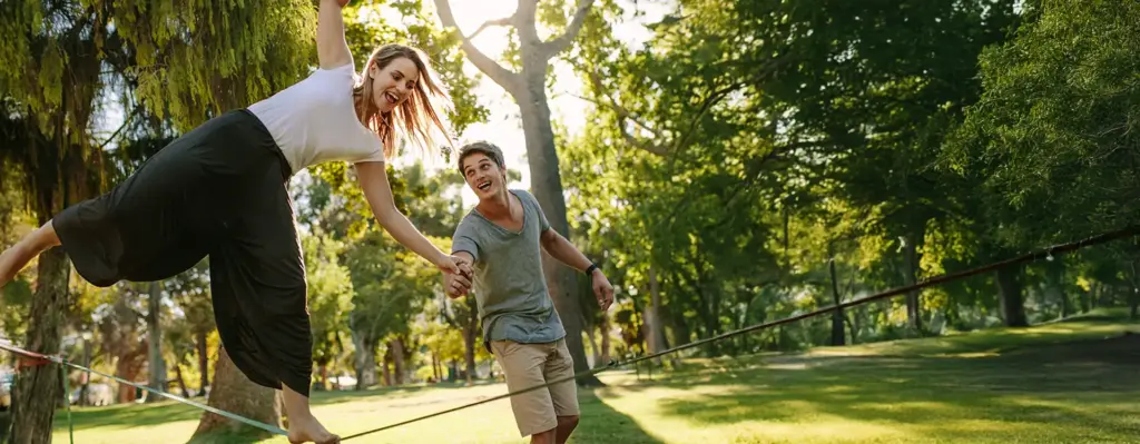 Junges Paar beim Slacklinen im Park – Frau balanciert auf dem Seil mit erhobenen Armen, Mann hält lachend ihre Hand.