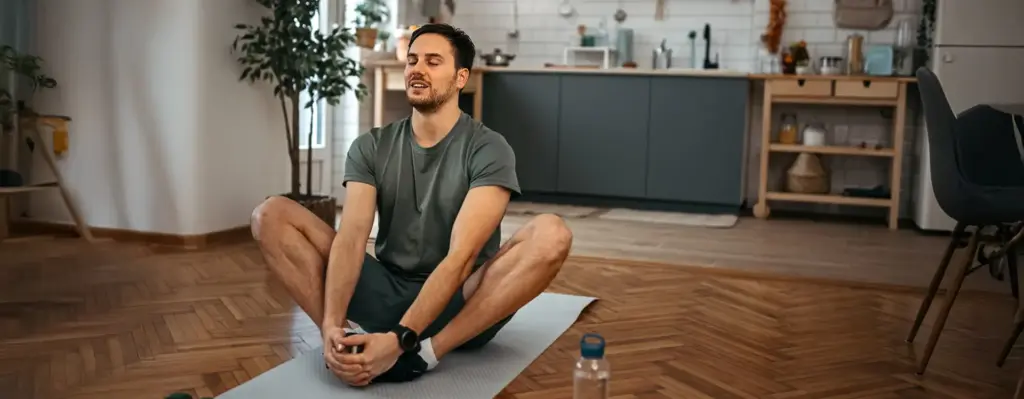 Junger Mann sitzt entspannt im Schneidersitz auf einer Yogamatte zuhause – Augen geschlossen, Wasserflasche neben ihm, nach dem Sport.