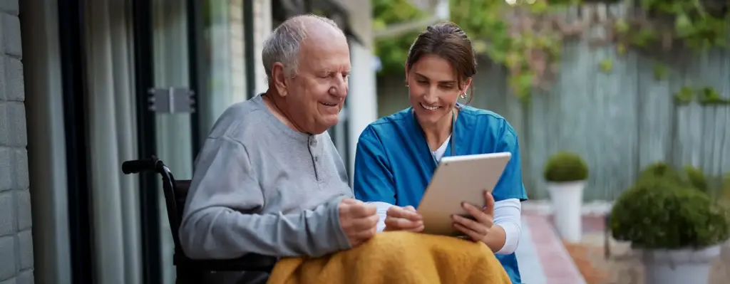 Pflegerin zeigt einem älteren Herrn etwas auf einem Tablet – beide lächeln, sitzen gemeinsam draußen, fürsorgliche Betreuung.