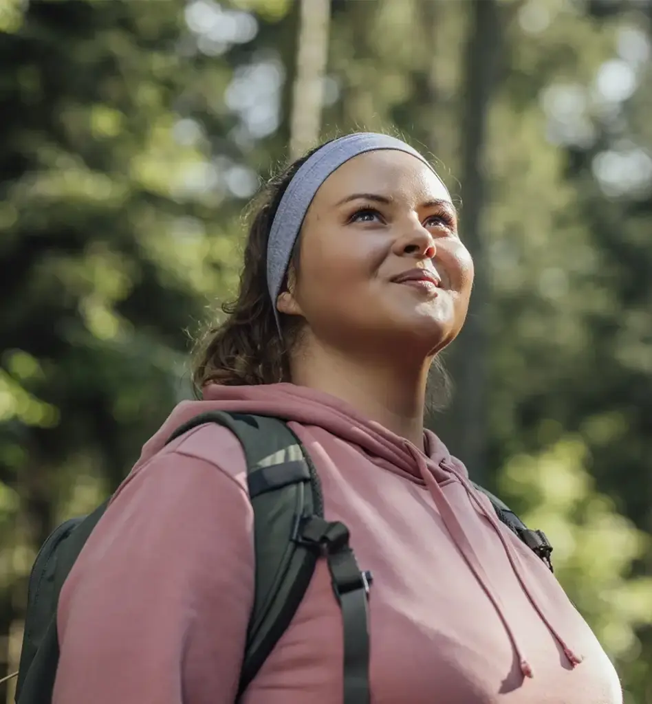 Frau mit rosa Hoodie, Stirnband und Rucksack blickt lächelnd nach oben – Nahaufnahme beim Waldspaziergang im Gegenlicht.