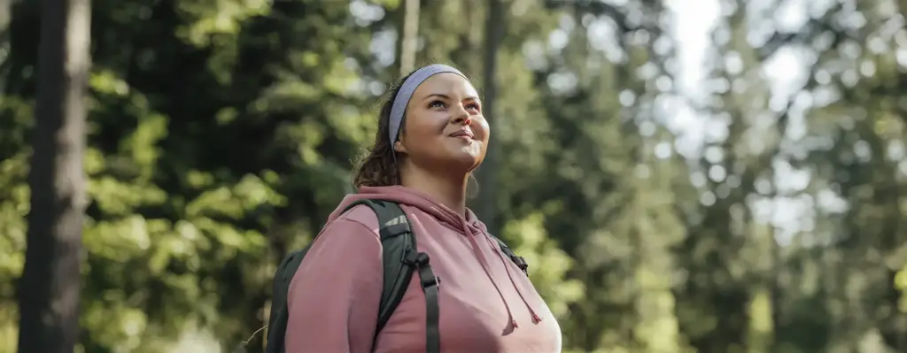Frau mit rosa Hoodie, Stirnband und Rucksack blickt lächelnd nach oben – Nahaufnahme beim Waldspaziergang im Gegenlicht.