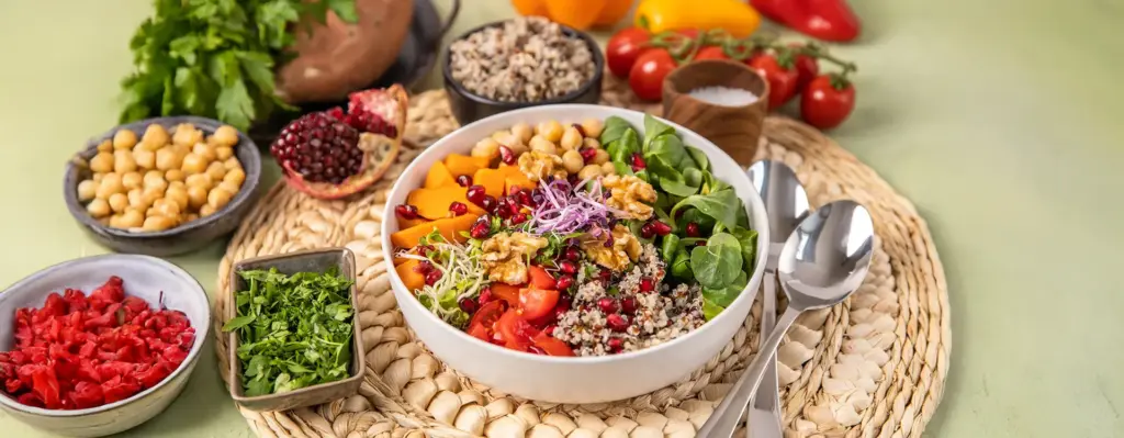 Bunte vegane Bowl mit Quinoa, Kichererbsen, Granatapfelkernen, Walnüssen, Tomaten und Feldsalat in einer weißen Schüssel – frisches Gemüse und Früchte im Hintergrund.