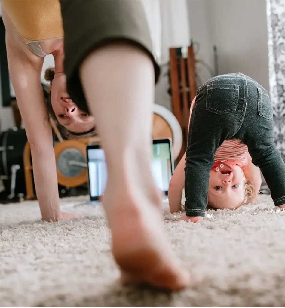 Mutter und kleines Kind machen gemeinsam Turnübungen auf dem Teppich – beide bücken sich kopfüber nach vorne und lachen sich an.