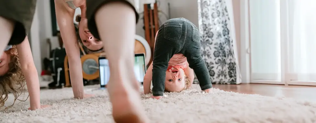Mutter und kleines Kind machen gemeinsam Turnübungen auf dem Teppich – beide bücken sich kopfüber nach vorne und lachen sich an.