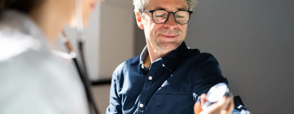 Ein Mann bekommt Blutdruck gemessen. Im Vordergrund ist eine medizinische Fachangestellt zu sehen.