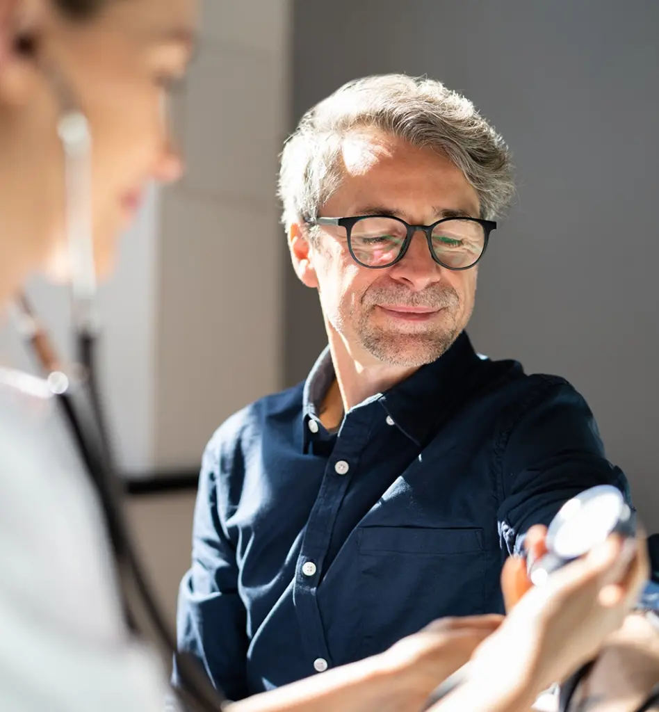 Ein Mann bekommt Blutdruck gemessen. Im Vordergrund ist eine medizinische Fachangestellt zu sehen.