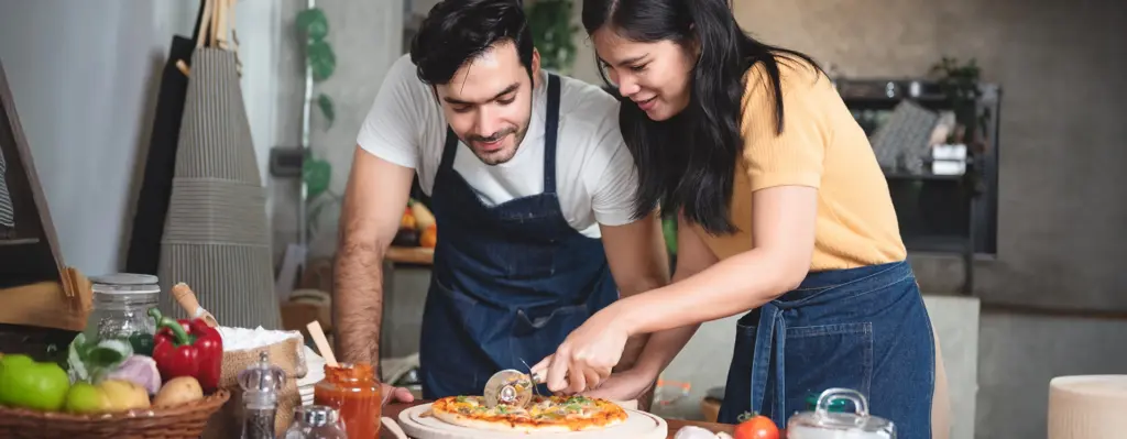 Eine Frau und ein Mann stehen an einem Tisch und die Frau schneidet eine Pizza mit einem Pizzaschneider.