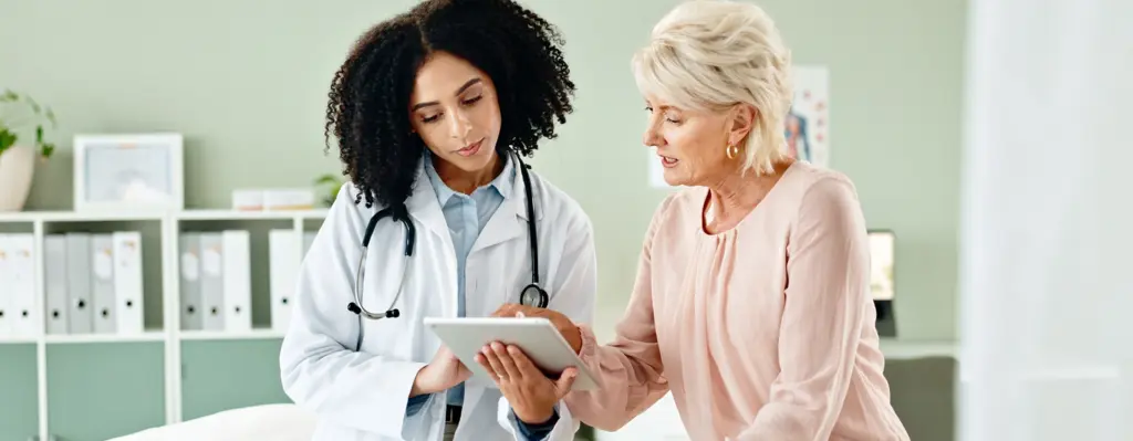 Eine Frau sitzt auf einer Untersuchungsliege. Neben ihr steht eine Ärztin mit einem Tablet und zeigt der Patientin etwas.