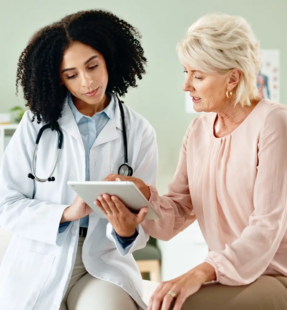 Eine Frau sitzt auf einer Untersuchungsliege. Neben ihr steht eine Ärztin mit einem Tablet und zeigt der Patientin etwas.