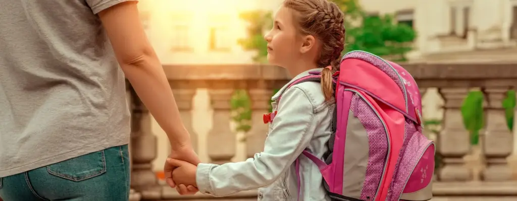 Ein Kind mit Schulrucksack hält die Hand einer erwachsenen Person und blickt diese an.