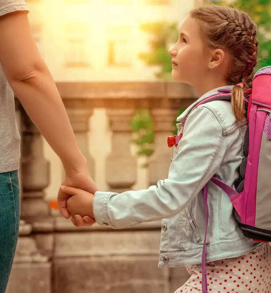 Ein Kind mit Schulrucksack hält die Hand einer erwachsenen Person und blickt diese an.