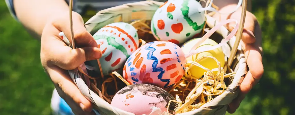 Man sieht ein Osternetz mit bunt angemalten Eiern. Das Nest wird von Kinderhänden gehalten.