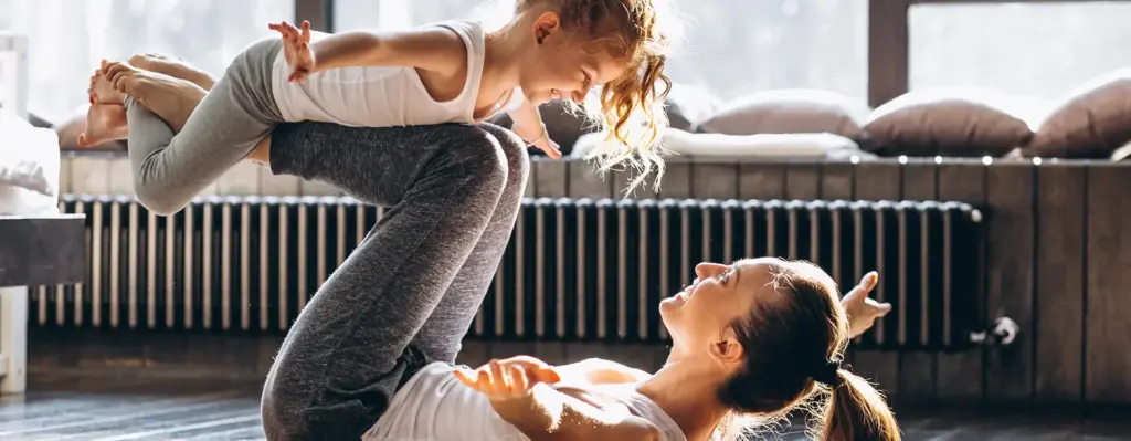Eine Frau liegt auf einer Sportmatte mit angewinkelten Beinen und balanciert ein Mädchen auf ihren Beinen.