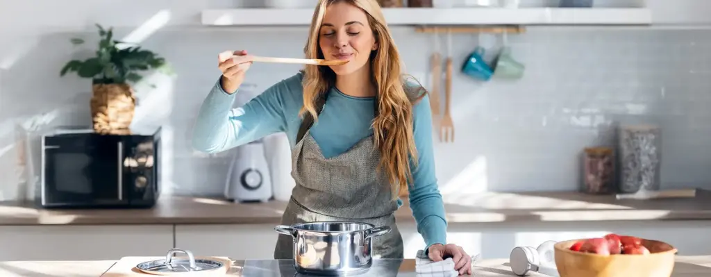 Eine Frau steht in der Küche mit Schürze und probiert von einem Kochlöffel.