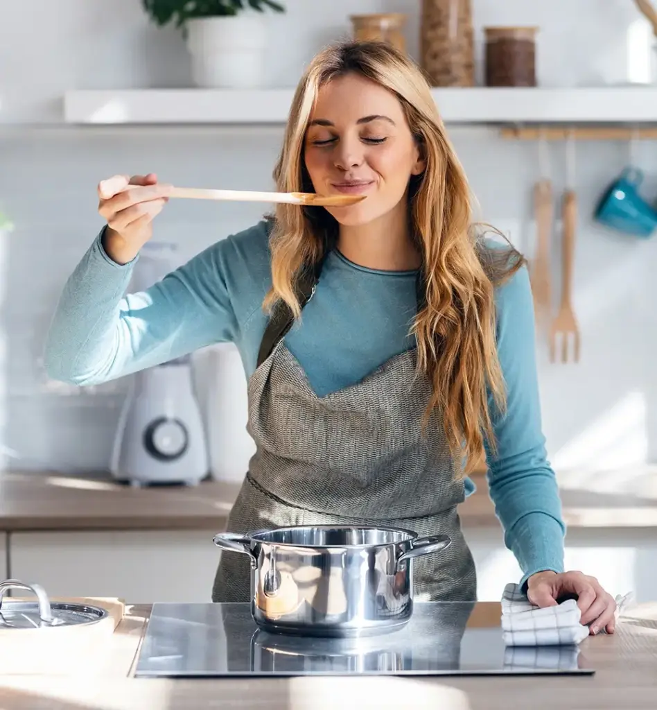 Eine Frau steht in der Küche mit Schürze und probiert von einem Kochlöffel.