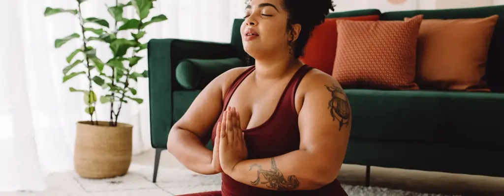 Eine Frau in Sportkleidung sitzt vor einem Sofa auf dem Boden im Schneidersitz und hat die Hände vor der Brust in Gebetshaltung gefaltet. Das Bild wird im Bereich "7Mind App – weil Balance wichtig ist!" verwendet.