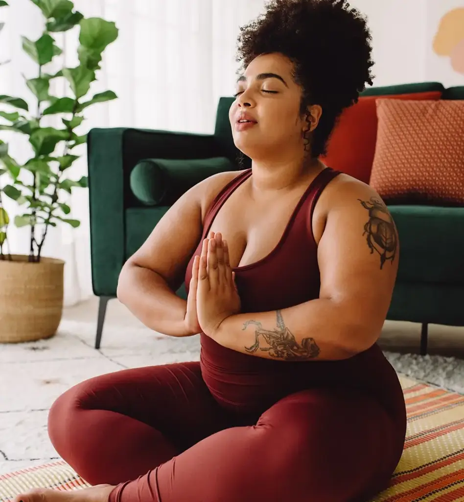 Eine Frau in Sportkleidung sitzt vor einem Sofa auf dem Boden im Schneidersitz und hat die Hände vor der Brust in Gebetshaltung gefaltet. Das Bild wird im Bereich "7Mind App – weil Balance wichtig ist!" verwendet.