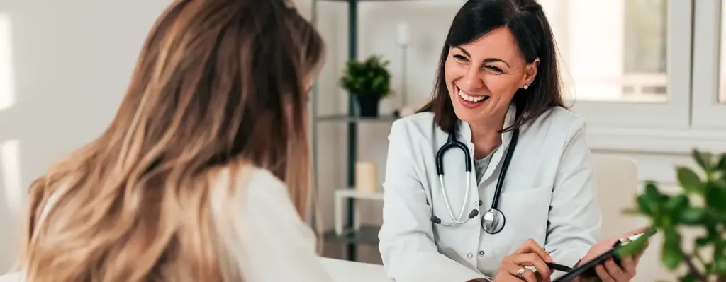 Eine Ärztin sitzt am Schreibtisch und zeigt einer Patientin, die gegenüber sitzt etwas auf einem Klemmbrett. Das Bild wird im Bereich "Ärztliche Behandlung" verwendet.