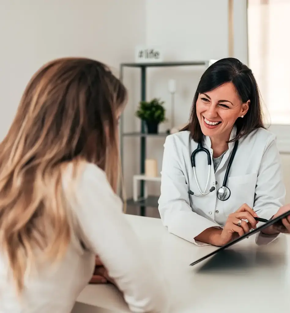 Eine Ärztin sitzt am Schreibtisch und zeigt einer Patientin, die gegenüber sitzt etwas auf einem Klemmbrett. Das Bild wird im Bereich "Ärztliche Behandlung" verwendet.