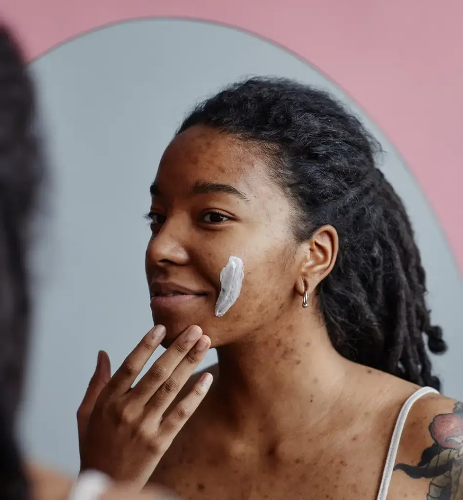 Eine jüngere Frau mit Hautirritationen schmiert sich Creme auf die Wange und blickt in einen Spiegel. Das Bild wird im Bereich "Akne - Behandlung mit Lichttherapie" verwendet.