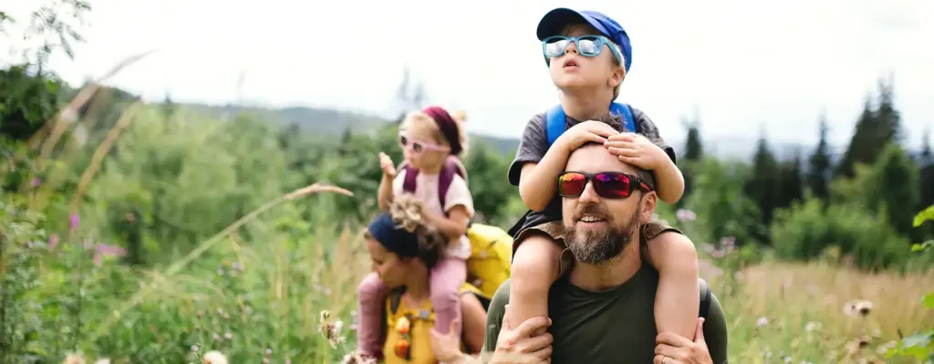 Ein Mann hat einen kleinen jungen auf en Schultern sitzen. Sie tragen beide eine Sonnenbrille und der Junge auch eine Cap. Im Hintergrund ist eine Frau mit einem Mädchen auf den Schultern zu sehen. Die Familie ist in der Natur unterwegs. Das Bild wird im Bereich "Aktivbonus - jetzt holen!" verwendet.