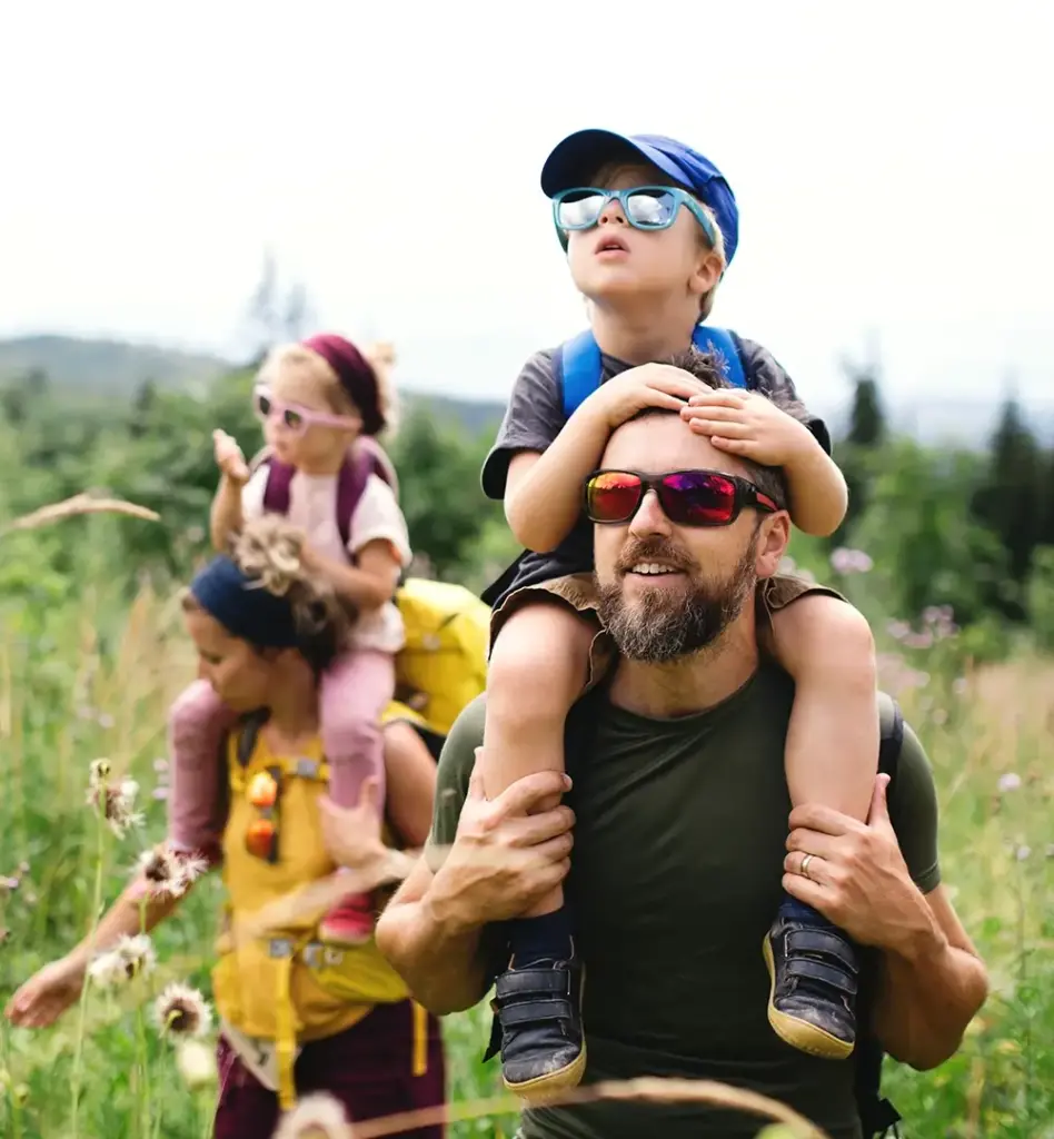 Ein Mann hat einen kleinen jungen auf en Schultern sitzen. Sie tragen beide eine Sonnenbrille und der Junge auch eine Cap. Im Hintergrund ist eine Frau mit einem Mädchen auf den Schultern zu sehen. Die Familie ist in der Natur unterwegs. Das Bild wird im Bereich "Aktivbonus - jetzt holen!" verwendet.