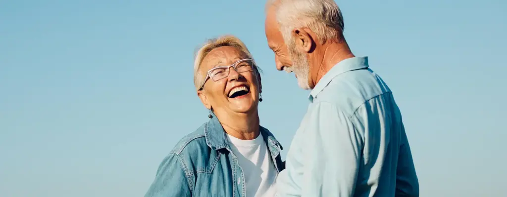 Eine ältere Frau lacht und steht einem älteren Mann gegenüber. Sie stehen im Freien. Das Bild wird im Bereich "Aktive Gesundheitsreisen" verwendet.