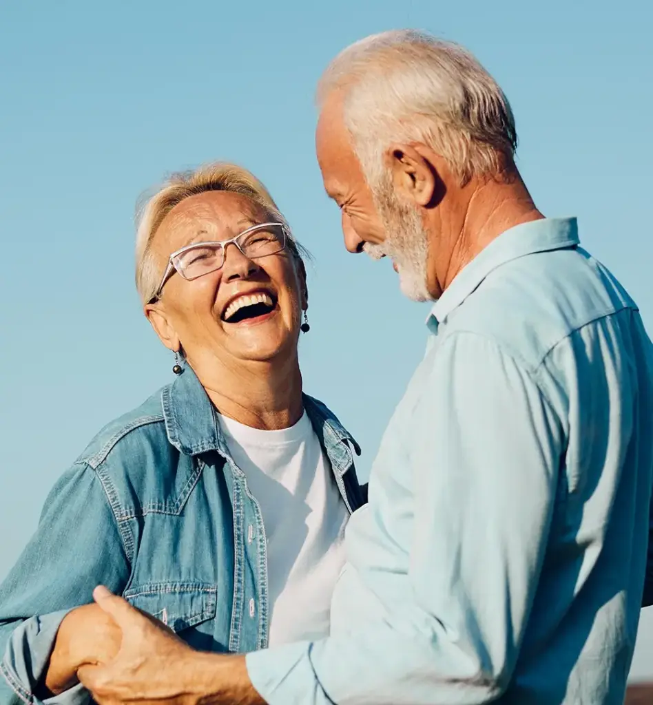 Eine ältere Frau lacht und steht einem älteren Mann gegenüber. Sie halten ihre Hände. Sie stehen im Freien. Das Bild wird im Bereich "Aktive Gesundheitsreisen" verwendet.