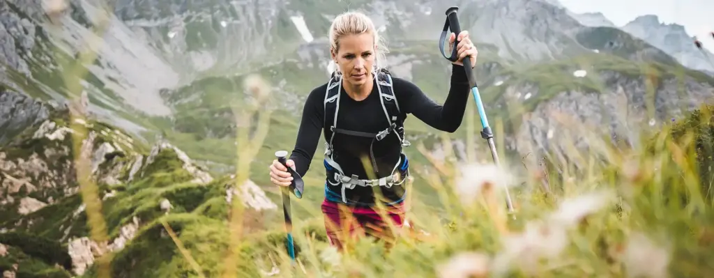 Eine junge Frau ist in den Bergen unterwegs. Sie hält zwei Wanderstöcke in der Hand und hat einen Rucksack auf dem Rücken. Das Bild wird im Bereich "Aktivkuren in Europa" verwendet.