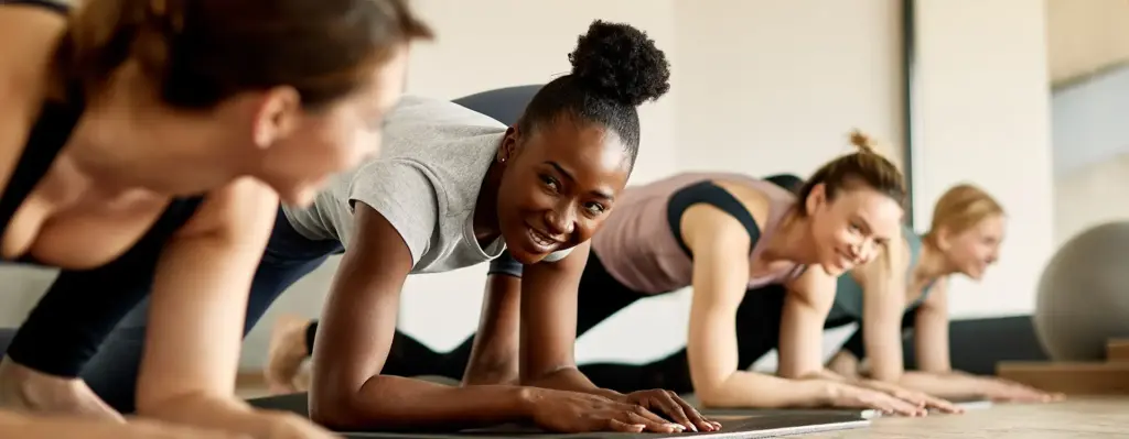 Drei Frauen dehnen sich auf Fitnessmatten, in dem sie eins ihrer Knie nach vorne ziehen und sich mit dem anderen Bein und den Armen in Liegestütz-Position befinden. Das Bild wird im Bereich "Aktivwoche" verwendet.