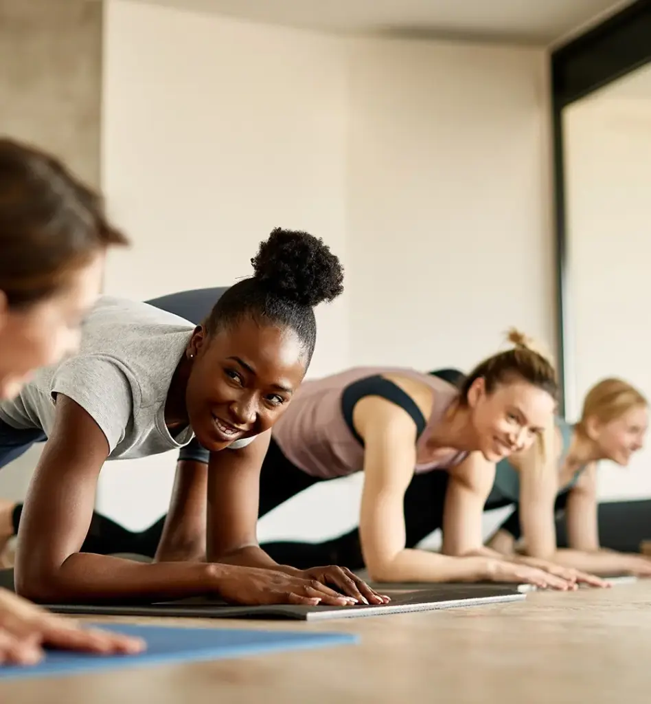 Drei Frauen dehnen sich auf Fitnessmatten, in dem sie eins ihrer Knie nach vorne ziehen und sich mit dem anderen Bein und den Armen in Liegestütz-Position befinden. Das Bild wird im Bereich "Aktivwoche" verwendet.