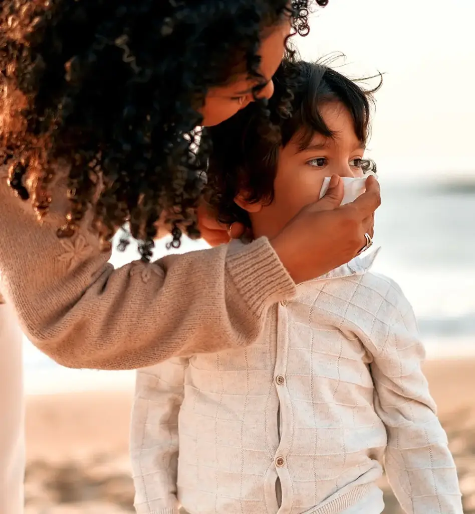 Eine Frau beugt sich zu einem kleinen Jungen runter und putzt ihm mit einem Taschentuch die Nase. Das Bild wird im Bereich "Auslands-Versicherungsschutz" verwendet.