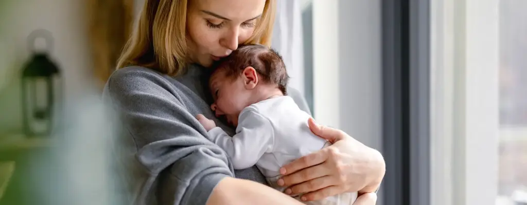 Eine Frau hält ihr Neugeborenes auf dem Arm und gibt ihm einen Kuss auf den Kopf. Das Bild wird im Bereich "BabyCare Nutrition - Ihre Ernährung für die gesunde Entwicklung Ihres Kindes" verwendet.